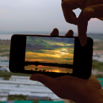 Image: Hands hold up a cellphone displaying a bright sunset photograph on the screen. The cellphone is lined up to match the angle from which the photo was taken, almost aligning with the horizon. Beyond the frame of the cellphone—gray sky, shrubs, waste, tin sheets, and green tarpaulin roofs.