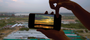 Image: Hands hold up a cellphone displaying a bright sunset photograph on the screen. The cellphone is lined up to match the angle from which the photo was taken, almost aligning with the horizon. Beyond the frame of the cellphone—gray sky, shrubs, waste, tin sheets, and green tarpaulin roofs.
