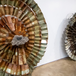 Image: Detail of a photograph of work by Naomi Wanjiku; three large spiraling works of folded metal rest on the floor against the white gallery wall.