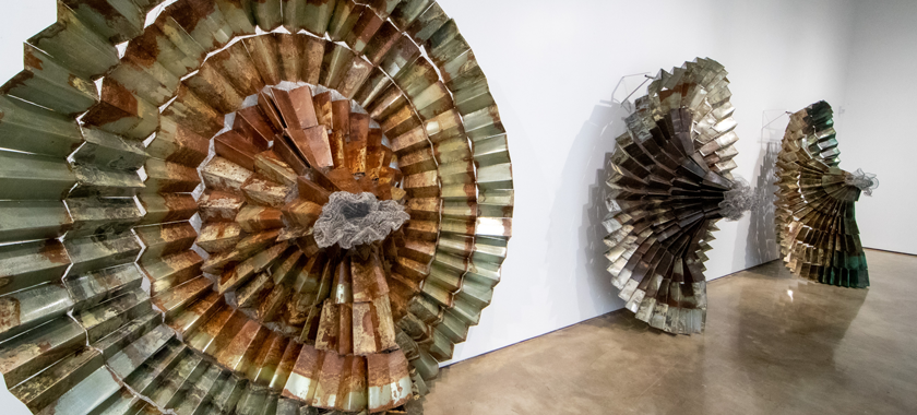 Image: Detail of a photograph of work by Naomi Wanjiku; three large spiraling works of folded metal rest on the floor against the white gallery wall.