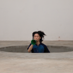 Image detail: Aki Sasamoto (Fellow in Interdisciplinary Work ’18), random memo random, 2016, Mixed-media performance/installation. There is a big circular hole dug into a concrete floor of an industrial space. The artist is jumping up and down in this hole. In the photo, you see her upper body above the ground and the rest is inside the hole, with floating hair that indicates her jumping motion. She holds a megaphone, into which she shouts.