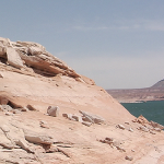 Image: A.K. Burns (Fellow in Interdisciplinary Works ’18), Video still from ‘A Smeary Spot’, 2015, HD video installation. In this video still we see a large pinkish land mass in the foreground, without vegetation. In the distance are other large desert rock formations and a body of water at the base of the foregrounded land mass. On the top of the foregrounded mass is a performer seen from a distance, waving a large piece of silver reflective material in the air. Presumably they are mid dance.