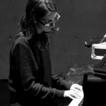 Image: detail of a black and white photograph of composer Teodora Stepančić, who is seated at a piano or keyboard, head down as she concentrates on the keys. She has dark hair that grows past her shoulders, and wears plastic glasses frames and a long-sleeved top.