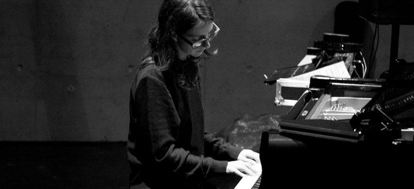 Image: detail of a black and white photograph of composer Teodora Stepančić, who is seated at a piano or keyboard, head down as she concentrates on the keys. She has dark hair that grows past her shoulders, and wears plastic glasses frames and a long-sleeved top.