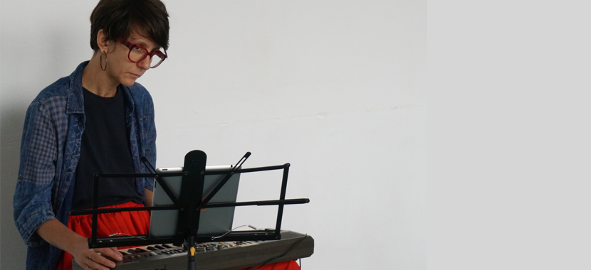 Image: Detail of a color photo of composer Teodora Stepančić, seated at a keyboard with a music stand in front. Stepančić wears plastic-framed glasses, hoop earrings, and has short hair that frames her face. She is wearing a black tee-shirt with a denim-colored button down top and a bright red skirt.