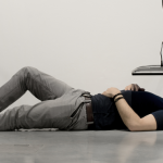 Image Detail: an individual is laying on their back on a smooth concrete floor, their head under a TV monitor that is mounted to the wall, screen facing down towards their face.