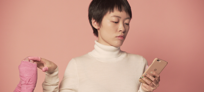 Image Detail: Photograph of an individual wearing a white turtleneck, eyes gazing down at their sleek mobile phone and situated against a pink backdrop; they hold one hand out, touching another hand that is wrapped in pink bandages.