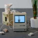 An exhibition view shows a group of objects assembled on the concrete floor. Two 1980’s era Macintosh computers are surrounded by a plaster cast polyhedron, stones, wires, cast resin Apple mice, a partial 3D print of the bust of Nefertiti, and a plastic plant. The computer screens display black and white images of other plastic plants that can be purchased on the internet.