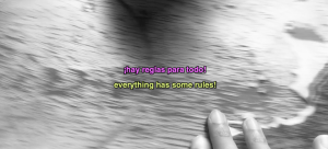 Still frame from a black and white video: fingers smear water over a wooden surface. Subtitles in the middle of the frame read in Spanish (pink text): "¡hay regals para todo!" and in English (yellow text): "everything has some rules!"