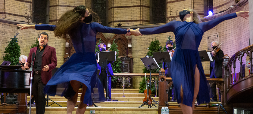 Two dancers in blue dresses grasp wrists with one arm outstretched, a singer in a burgundy jacket and musicians behind them in a church-like setting.