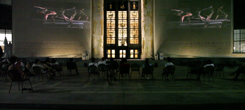 A photograph of the facade of the Brooklyn Public Library, a crowd seated on the plaza looking at two videos of dancers projected on the building.