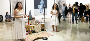Two performers in white dresses stand at a makeshift altar in a gallery space, addressing a crowd that is gathered around them.