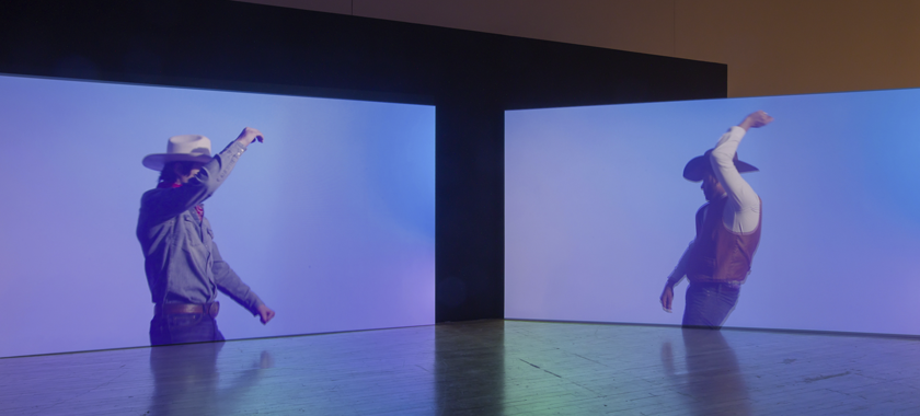 The image shows two projection screens that are slightly angled towards each other in an art gallery. Each screen shows a figure dressed in cowboy clothing while in front of a blue/pink/purple background. The two figures are facing each other, and are caught mid-movement, with their arms in the air.
