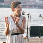 Régine M. Roumain stands at a microphone, hands mid-clap, addressing a crowd with the East River behind her
