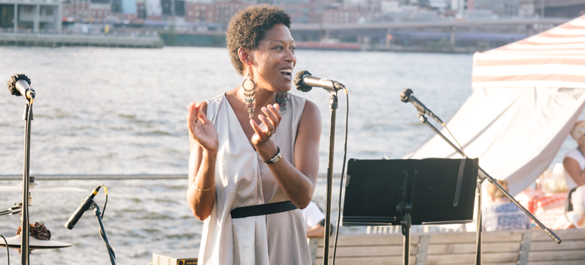 Régine M. Roumain stands at a microphone, hands mid-clap, addressing a crowd with the East River behind her