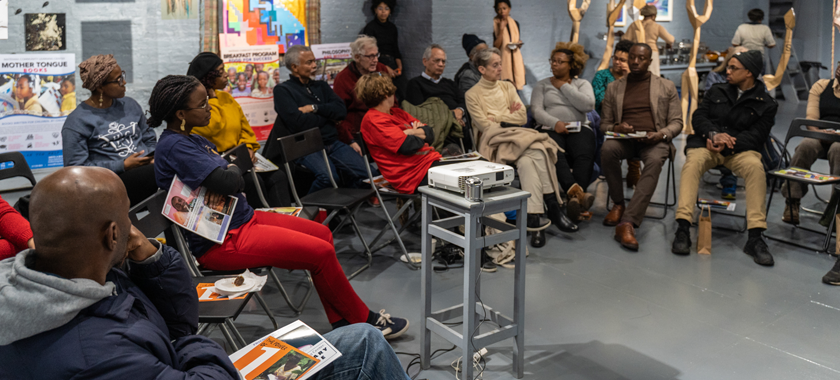 Individuals sit in chairs that have been set up in a circle, a project at the center and artworks in the background