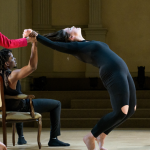 Hilary in a fuschia dress is standing facing back, slightly covering Rahmus in a black vest and pants seated in a chair with his legs wide. Hilary and Rahmus clasp Kristina’s wrist with a single hand to the left, as she in a black leotard performs a deep body bend profile. Briana in a nude blouse and black skirt is seated on the floor to the right of the others looking up at them.