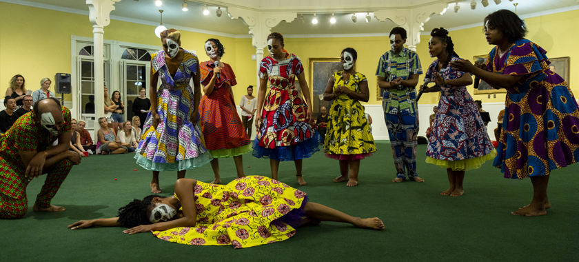 Eight performers look down on a dancer that is passed out on the ground in the large ballroom with the green carpet. They look concerned for her. One male dancer kneels on one knee above the dancer that lays on the ground. They are all wearing tailored, brightly colored West African print western style dresses. The women have brightly colored petticoats underneath their dresses.