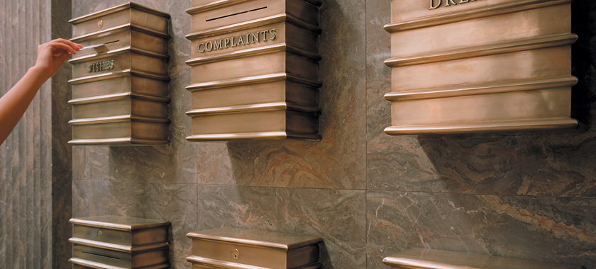 Photo detail of an arm outstretched, hand reaching towards a slot in a bronze-colored metal box on a marbled wall. The box is labeled "Wishes," and there are other boxes installed around it with labels including "Complaints" and "Dreams."