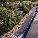 Aerial shot of a widened sidewalk that is entirely covered with a wavy pattern consisting of red, white, blue, yellow, and dark grey color ribbons made of dyed concrete with glass aggregates. Along the edges of the path, some ribbons are partly lifted to create small mounds of ambiguous shapes. The mounds also incorporate planters with lilyturf planting. People walk and bike on the path oftentimes following the wavy pattern; kids on their scooters and skateboards ride over the mounds.