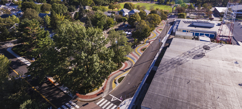 Aerial shot of a widened sidewalk that is entirely covered with a wavy pattern consisting of red, white, blue, yellow, and dark grey color ribbons made of dyed concrete with glass aggregates. Along the edges of the path, some ribbons are partly lifted to create small mounds of ambiguous shapes. The mounds also incorporate planters with lilyturf planting. People walk and bike on the path oftentimes following the wavy pattern; kids on their scooters and skateboards ride over the mounds.