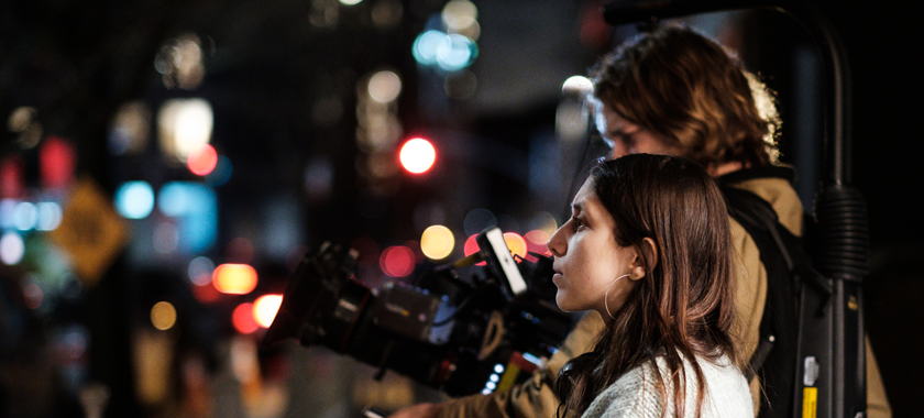 Two people stand together in a night city bright with lights. They face to the side. One person is holding a large professional camera.
