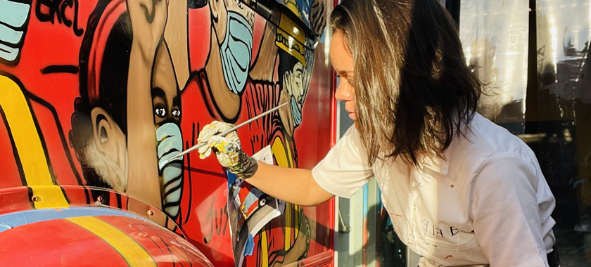 Lina Montoya painting a mural, focusing on the blue of a surgical mask over the face of an essential worker