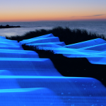 Ribbons of electric blue light cascade over the coastline as the sun sets.