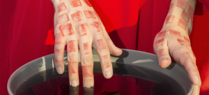 A hand covered in small, square red tattoos dips fingers into a bowl of dark water.