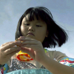 Yuki, a young Japanese-American girl, stands on the shore at Baby Makapuu in Oahu, examining a small shell.