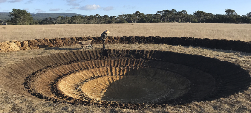 A dry grassy field under a blue sky. A crater stretches the width of the image and at the far side, the artist is seen digging with a shovel.