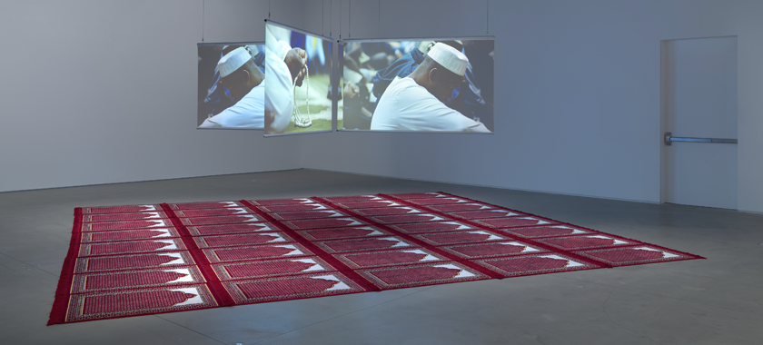 Floating 4-channel installation in a gallery space with red prayer mats below on the ground.