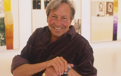 Image: A man, Robert Rauschenberg, is sitting in a white gallery-like setting in front of his own artworks. His hands are clasped together as he smiles into the camera.