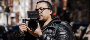 Photo of an individual wearing glasses, all black, and a backpack holding a film camera o a city street