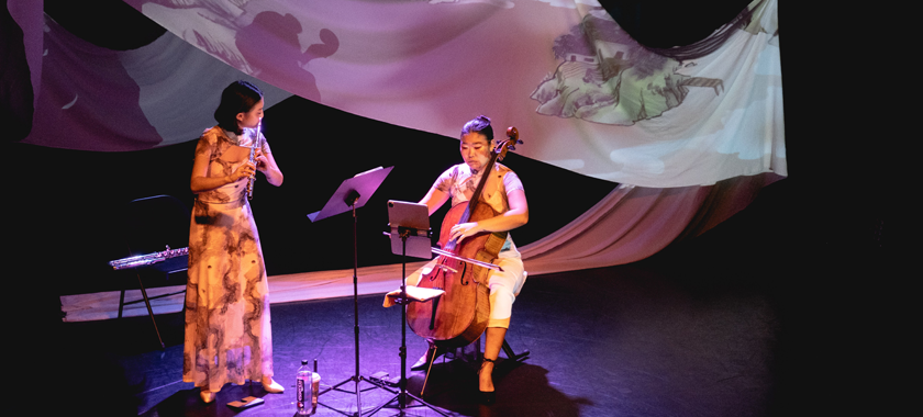 Two musicians--one playing the flute, the other playing the cello, perform on a darkened stage, fabric draped behind them with projections over top