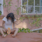 Detail of a video still of a woman wearing white, legs slightly open with a papaya in between
