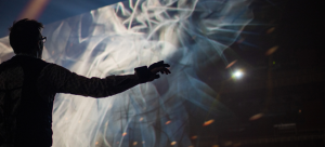 Male performer with gesture sensor glove is drawing graphics in the air, and the resulting scrim of projected graphics is shown, behind the figure, within the blackbox theater.