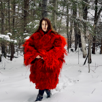 A woman stands in a snow-blanketed forested area, wearing a red Hunia, a traditional shepherd's wool raggedy coat