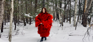 A woman stands in a snow-blanketed forested area, wearing a red Hunia, a traditional shepherd's wool raggedy coat