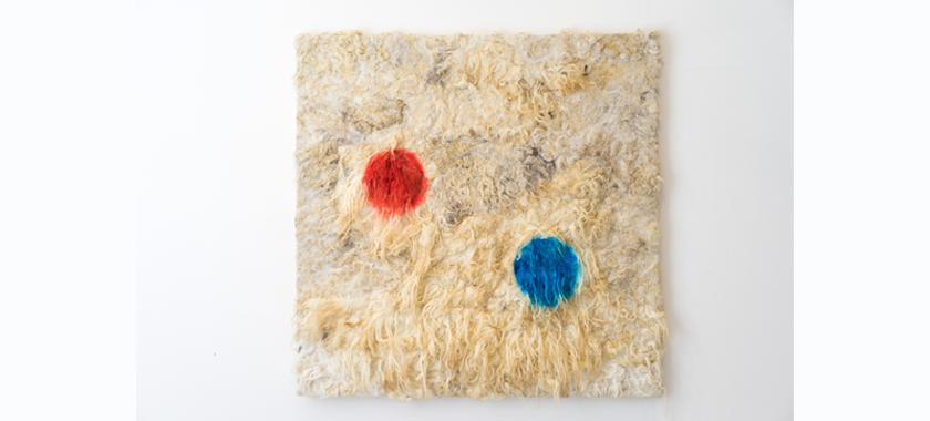 A square artwork of wool with one small red circle and one small blue circle painted on top of the natural fibre
