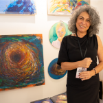 An artist in a black dress smiles at the camera against the backdrop of their artwork on display at Superfine