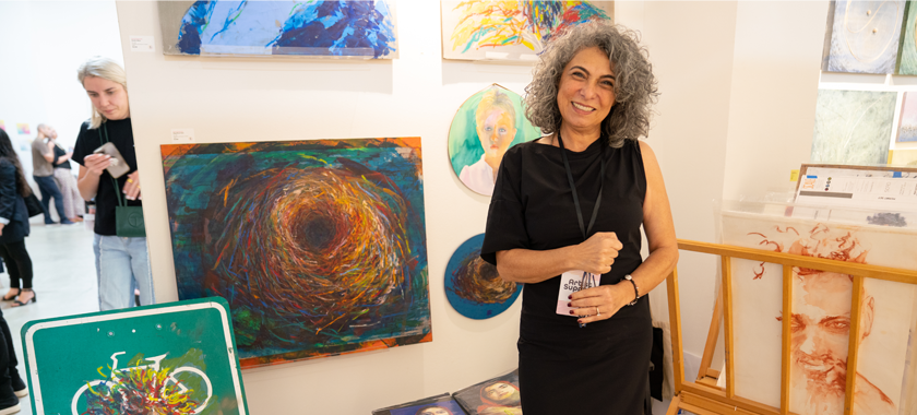 An artist in a black dress smiles at the camera against the backdrop of their artwork on display at Superfine
