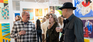 Three individuals stand at an art fair booth in conversation