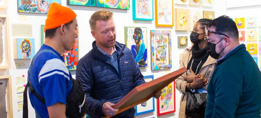 Four art fair attendees, three together and looking at a larger artwork with smaller artworks hung on the walls behind them