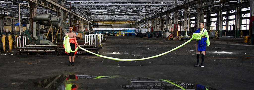 Two women stand in an abandoned industrial warehouse, connected by a chord of fabric.