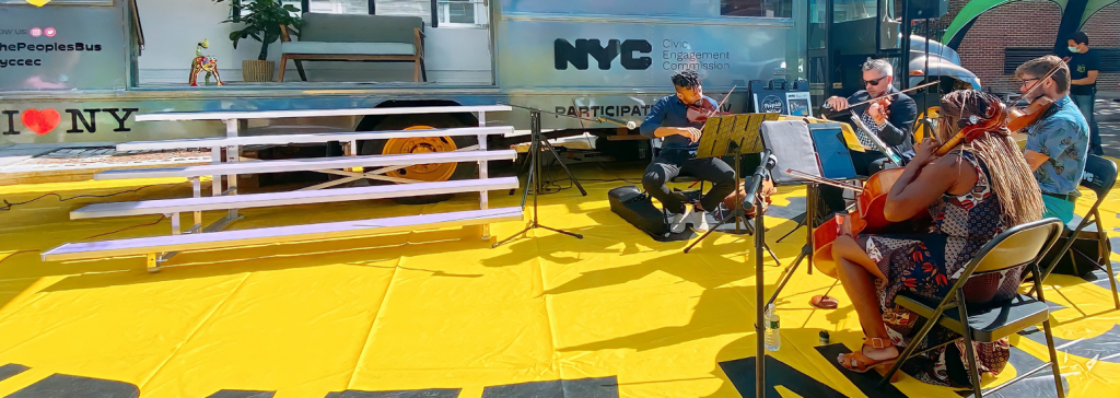 A group of 4 performers sit around a silver trailer. There is a yellow cover on the ground below them.