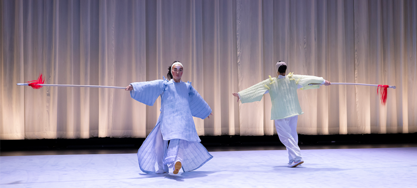 Two performers on a stage, one arm extended with a long pole with bright red threads coming off the end.