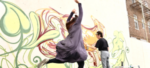 This picture depicts one dancer jumping with her arms in the air, while behind the dancer a painter is painting an image onto a large concrete wall using vibrant colors.