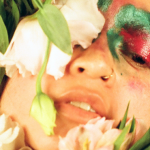 Close up of the artist dressed in a flower costume. Her face has pink and green make up and she is holding flowers in front of her face.