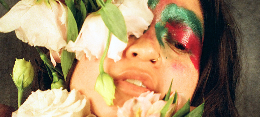 Close up of the artist dressed in a flower costume. Her face has pink and green make up and she is holding flowers in front of her face.